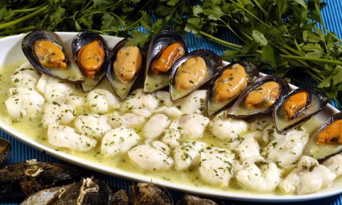 Una generosa fuente blanca repleta de carrilleras de rape (o cocochas) cocinadas en una brillante salsa verde con abundante perejil picado. El plato está elegantemente decorado con una hilera de mejillones al vapor en su concha, dispuestos de forma radial. La presentación, sobre un mantel azul y con ramos de perejil fresco al fondo, resalta la frescura y el carácter tradicional de este guiso marinero.