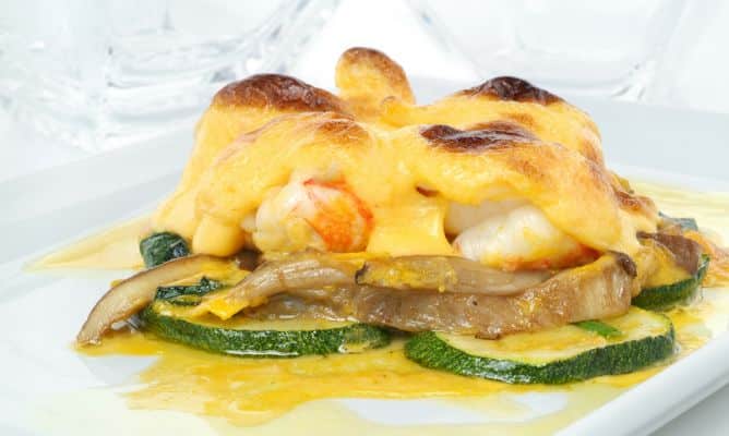 Una presentación gourmet de cigalas peladas y gratinadas con una muselina o salsa holandesa dorada al horno. El marisco se apoya sobre una base de rodajas de calabacín a la plancha y setas salteadas, todo ello bañado en una reducción de su propio jugo. El plato destaca por el contraste de texturas y el intenso color dorado del gratinado, servido en una vajilla blanca minimalista.