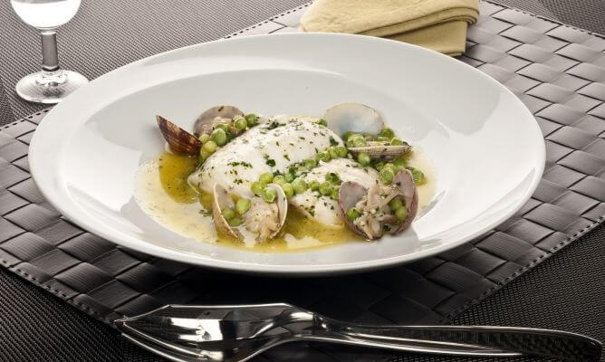 Un lomo de merluza perfectamente cocinado en una clásica salsa verde, servido en un elegante plato hondo blanco sobre un mantel individual trenzado de color negro. El plato está generosamente acompañado de almejas frescas abiertas y guisantes tiernos, todo espolvoreado con perejil picado. La cuidada presentación incluye una copa de vino y cubiertos de diseño, resaltando la calidad de este plato tradicional de la gastronomía marinera.
