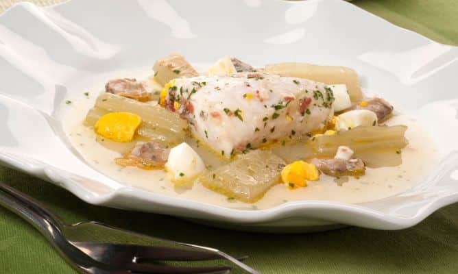 Un suculento lomo de merluza preparado en una suave salsa blanca, servido en un plato hondo con bordes ondulados. El pescado está acompañado de pencas de cardo tiernas, almejas y trozos de huevo cocido, aportando color y textura al conjunto. La presentación se completa con una lluvia de perejil picado y un toque de jamón, sobre un mantel verde que realza los tonos claros de esta receta tradicional y nutritiva.