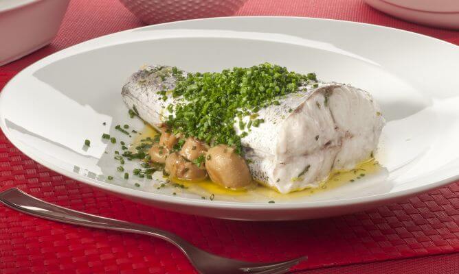 Un jugoso lomo de merluza cocinado al vapor o en papillote, manteniendo toda su textura y sabor natural. El pescado se presenta en un plato hondo blanco sobre una base de champiñones enteros salteados, cubierto generosamente con una capa de cebollino fresco picado. La presentación se completa con un chorrito de aceite de oliva virgen sobre un mantel individual de color rojo intenso, resaltando una opción culinaria saludable y refinada.