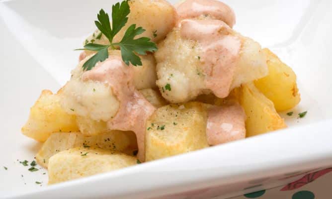Una ración de dados de rape empanados y fritos, servidos sobre una base de patatas doradas en un plato blanco hondo. El pescado está bañado con una suave salsa rosa o alioli de pimentón y decorado con una hoja de perejil fresco. Una presentación moderna y apetitosa que reinventa el concepto de las patatas bravas utilizando pescado blanco de calidad.