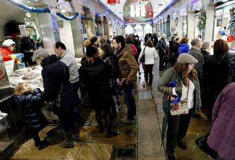 Una imagen vibrante del interior de un mercado tradicional lleno de clientes realizando sus compras para las fiestas. Los pasillos están concurridos por personas abrigadas, mientras los puestos de pescado fresco lucen decoraciones típicas como guirnaldas y luces azules. La escena refleja el bullicio característico de los días previos a Nochevieja, con consumidores buscando los mejores productos marinos para sus cenas de celebración.
