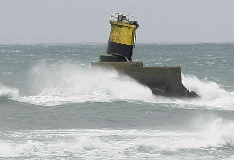 Una imagen de un faro o baliza de señalización marítima de colores amarillo y negro, situada sobre una base de hormigón en medio del mar. La estructura resiste el embate de un fuerte oleaje, con las olas rompiendo con fuerza y generando espuma blanca a su alrededor. El cielo gris y el mar picado reflejan un día de temporal en la costa, capturando la dureza del entorno marino.