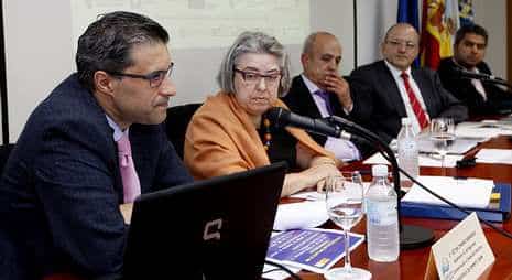 Una mesa redonda de expertos y representantes institucionales durante una jornada técnica o rueda de prensa. En primer plano, un ponente se dirige a la audiencia junto a un ordenador portátil, mientras el resto de los participantes escuchan con atención. El acto, que cuenta con carpetas de información y micrófonos, se desarrolla en un entorno formal con la presencia de banderas institucionales al fondo.