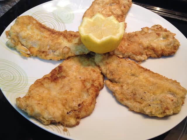 Bacaladilla rebozada con limón Cuatro filetes de bacaladilla rebozada en huevo y harina, fritos hasta alcanzar un tono dorado uniforme. Los lomos están dispuestos en forma de abanico en un plato blanco con detalles circulares verdes, coronados en el centro por un limón cortado en zigzag. Una presentación clásica de la cocina casera que resalta la sencillez y el sabor del pescado blanco.