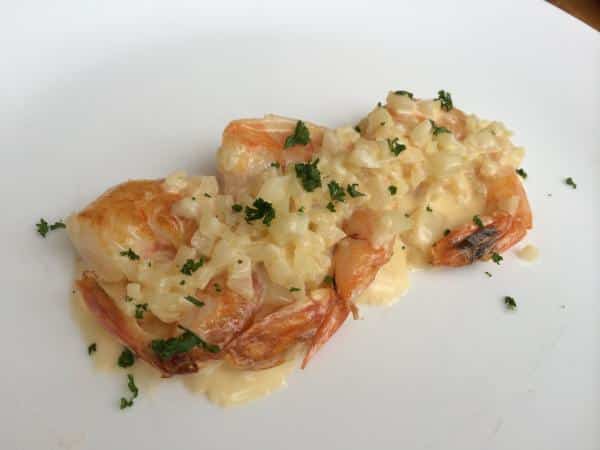 Una ración de gambas salteadas servidas en un plato blanco, cubiertas con una rica salsa de crema y trocitos de cebolla o ajo picado. El plato está decorado con perejil fresco esparcido por encima, ofreciendo una presentación elegante y cremosa que resalta el color rosado del marisco.