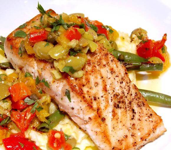 Un lomo de cachucho (o pargo) perfectamente marcado a la plancha, con la piel dorada y espolvoreado con pimienta negra. El pescado se presenta sobre una cama de puré y judías verdes, cubierto con una colorida vinagreta de aceitunas verdes, pimientos rojos y amarillos picados y perejil fresco, creando una combinación de sabores mediterráneos muy vibrante.