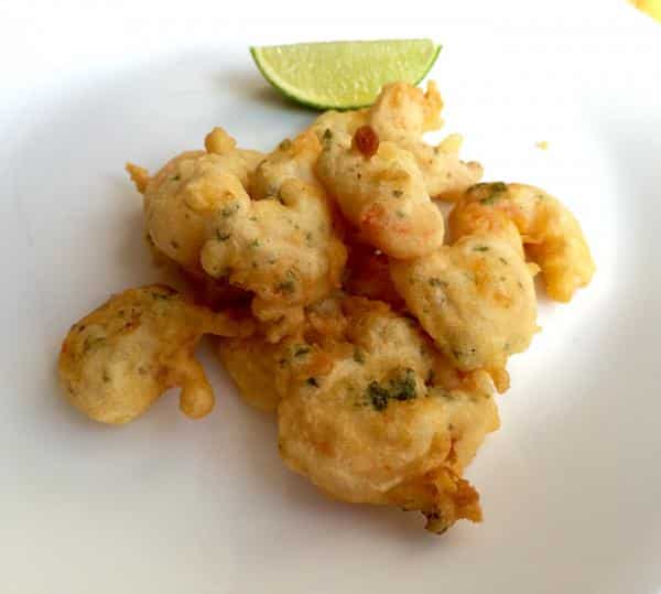 Una ración de buñuelos de gambas dorados y crujientes, servidos sobre un plato blanco liso. La masa, que deja ver pequeños trozos de perejil, tiene una textura aireada y se presenta acompañada de un gajo de lima fresca al fondo para aportar un toque cítrico al frito.