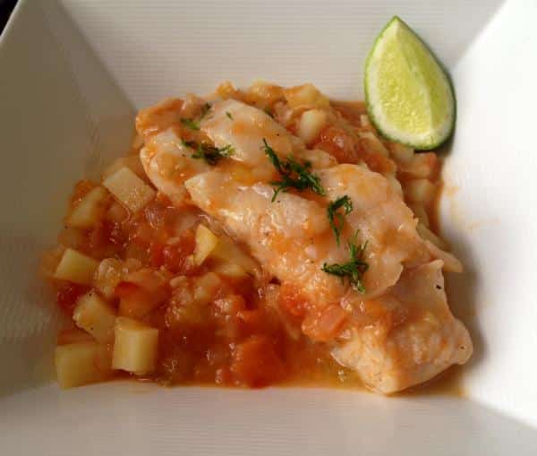 Un lomo de merluza jugoso cocinado a la cazuela, servido sobre una base de patatas en dados y un sofrito de tomate y cebolla. El plato está espolvoreado con eneldo fresco y se acompaña de un gajo de lima en el borde de un cuenco blanco cuadrado, ofreciendo una presentación sencilla y sabrosa.