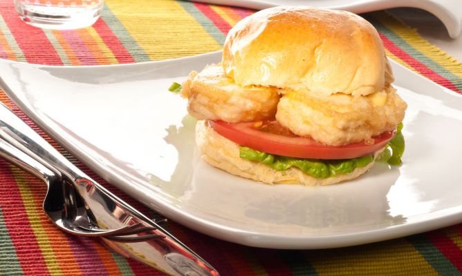 Una apetitosa mini hamburguesa de merluza servida en un plato blanco de diseño moderno. El relleno consiste en lomos de merluza suavemente rebozados, una rodaja de tomate natural y una hoja de lechuga fresca, todo dentro de un tierno pan de brioche brillante. Se presenta acompañada de cubiertos sobre un mantel de rayas coloridas, ideal para un menú infantil o un aperitivo original.