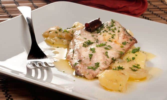 Un jugoso filete de jurel al horno servido en un plato blanco cuadrado, acompañado de patatas panadera tiernas. El pescado está aderezado con perejil fresco picado, láminas de ajo y una guindilla seca que aporta un toque sutil de picante. El conjunto se presenta con un tenedor al lado sobre un mantel de bambú, resaltando una receta saludable y tradicional.
