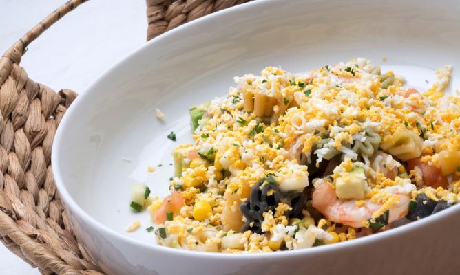 Una fresca ensalada de pasta con hélices tricolores, gambas peladas, maíz, tomate y calabacín, servida en un gran bol blanco dentro de un soporte de mimbre. El plato está generosamente cubierto con huevo cocido rallado y espolvoreado con perejil, ofreciendo una presentación colorida y saludable de inspiración mediterránea.