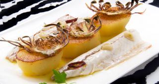 Un elegante plato de lomos de caballa perfectamente cocinados, acompañados de tres patatas fondant doradas. Cada patata está coronada con un crujiente chip de alcachofa frito, aportando una textura vertical al conjunto. El plato se termina con láminas de ajo, aros de guindilla y una base de aceite de oliva, presentado sobre un mantel de patrón geométrico en blanco y negro.