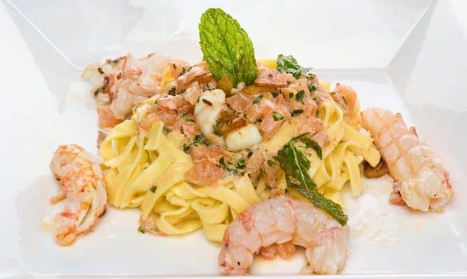 Un plato gourmet de tallarines coronado con colas de cigalas peladas y salteadas. La pasta está mezclada con una salsa ligera de tomate picado y hierbas frescas, decorada con una hoja de hierbabuena vertical en el centro. El conjunto se presenta en un plato blanco de líneas modernas, resaltando la frescura de los ingredientes marinos.