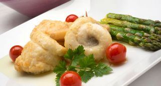 Dos lomos de gallo en escabeche enrollados de forma elegante y servidos en un plato blanco alargado. La presentación se completa con una guarnición de espárragos verdes a la plancha, varios tomates cherry rojos y brillantes, y una rama de perejil fresco sobre el pescado, todo sobre una base de aceite o salsa ligera.