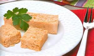 Tres porciones de pastel de cabracho (o tiñosu) servidas en un plato blanco con un suave relieve de puntos. El pastel, de textura esponjosa y color rosado anaranjado, está decorado con una hoja de perejil fresco y se presenta junto a un tenedor sobre un mantel rojo, destacando como un aperitivo clásico de la cocina marinera.