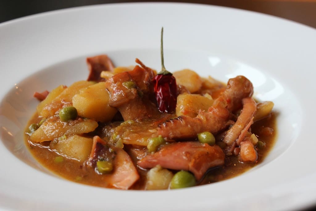 Un apetitoso guiso de pota (calamar gigante) servido en un plato hondo blanco. La receta incluye trozos tiernos de pota, patatas cocidas y guisantes en una salsa espesa y brillante. El conjunto está coronado con una guindilla roja seca en el centro, sugiriendo un toque picante y casero.