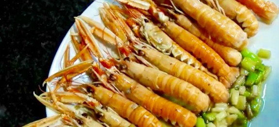 Una ración de cigalas cocinadas, dispuestas de forma ordenada en un plato blanco. Los crustáceos presentan un color anaranjado brillante y se acompañan de una guarnición de aceite, ajo y perejil picado en un lateral, resaltando la calidad del marisco fresco.