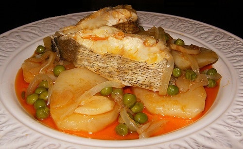 Un suculento plato de bertorella a la gallega servido en un plato hondo de cerámica blanca decorada. El pescado, con su carne blanca y piel brillante, se presenta sobre una cama de patatas cocidas, guisantes y cebolla, todo bañado en una tradicional ajada de color anaranjado.