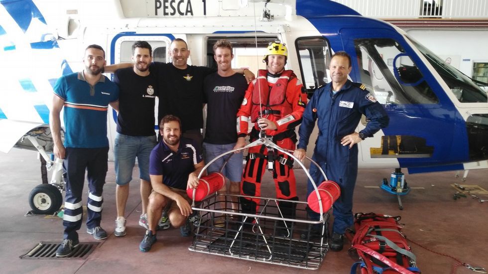 Siete hombres posan en un hangar junto al helicóptero de rescate Pesca 1. El grupo incluye personal de paisano y rescatistas uniformados, uno de ellos con traje de neopreno rojo y casco, situado junto a una cesta de salvamento marítimo. La aeronave, de colores blanco y azul, sirve de fondo para la fotografía del equipo.