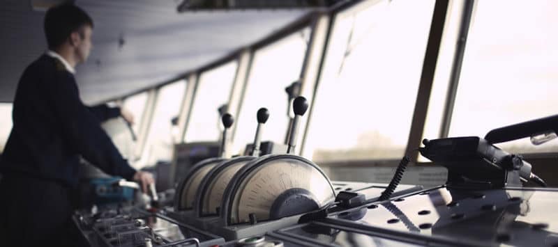 Vista del puente de mando de una embarcación con las palancas de control en primer plano y un oficial de marina de espaldas mirando hacia el horizonte.
