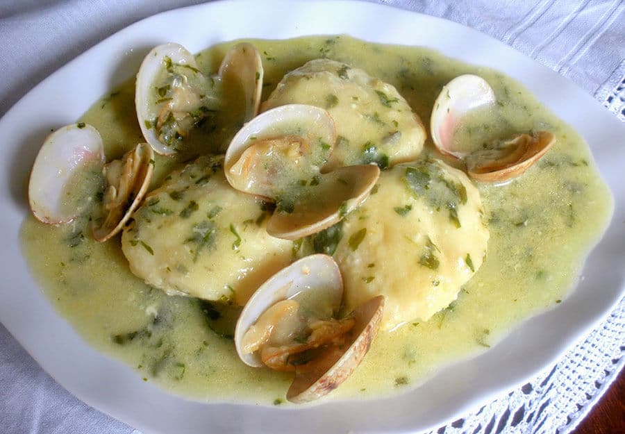 Medallones de rape en salsa verde con perejil, acompañados de almejas abiertas al vapor en un plato blanco.