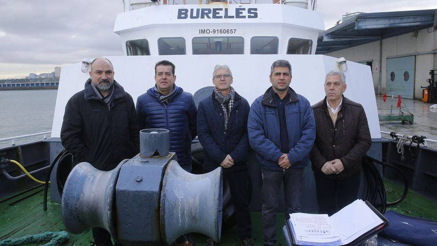 Cinco hombres posan frente a la cabina de un barco pesquero blanco con el nombre "BURELÉS" escrito en la parte superior. Están vestidos con ropa de abrigo y se encuentran en la cubierta de la embarcación, en lo que parece ser un puerto bajo un cielo nublado.
